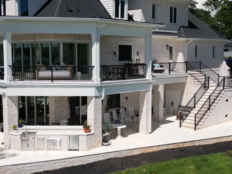 Rear elevation of a white lakefront home with stacked covered porches, black metal railings, and a built-in grill station on the lower patio