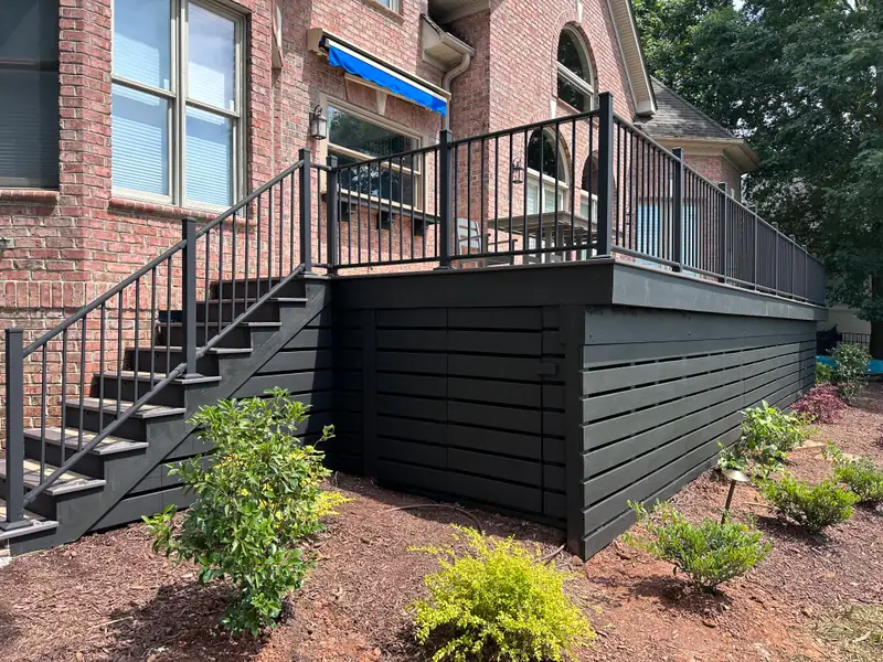 Brick home with an elevated deck, black aluminum railing, dark horizontal skirting, and a full-run stair to the yard