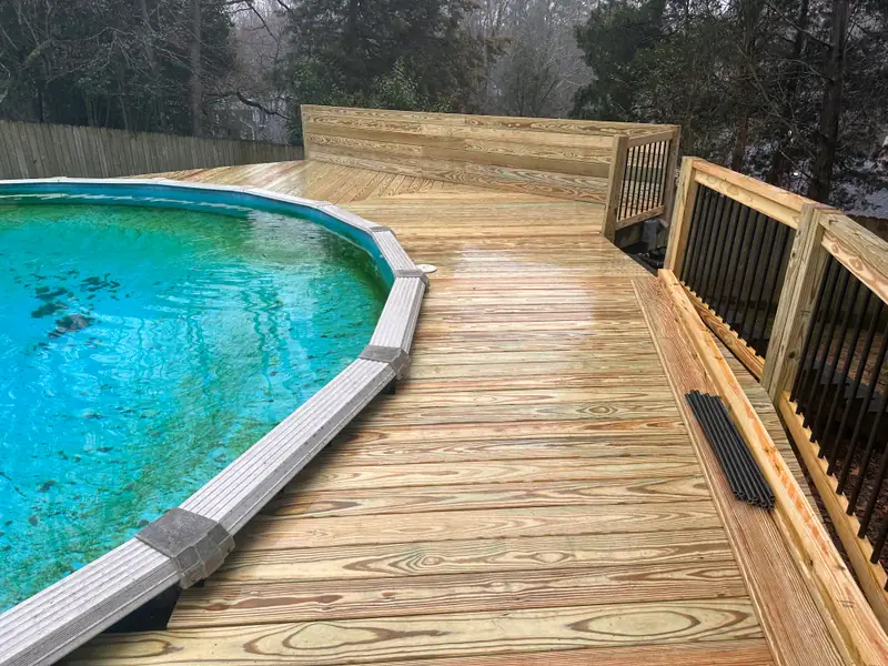 Pressure-treated wood deck built around an above-ground pool, with a wood rail and black balusters