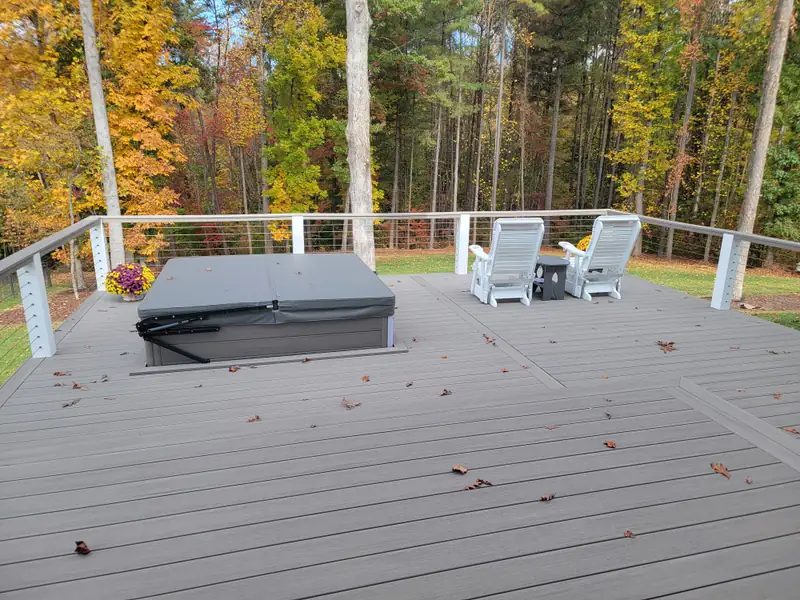 Gray composite deck with cable railing, a covered hot tub, and patio chairs facing autumn woods