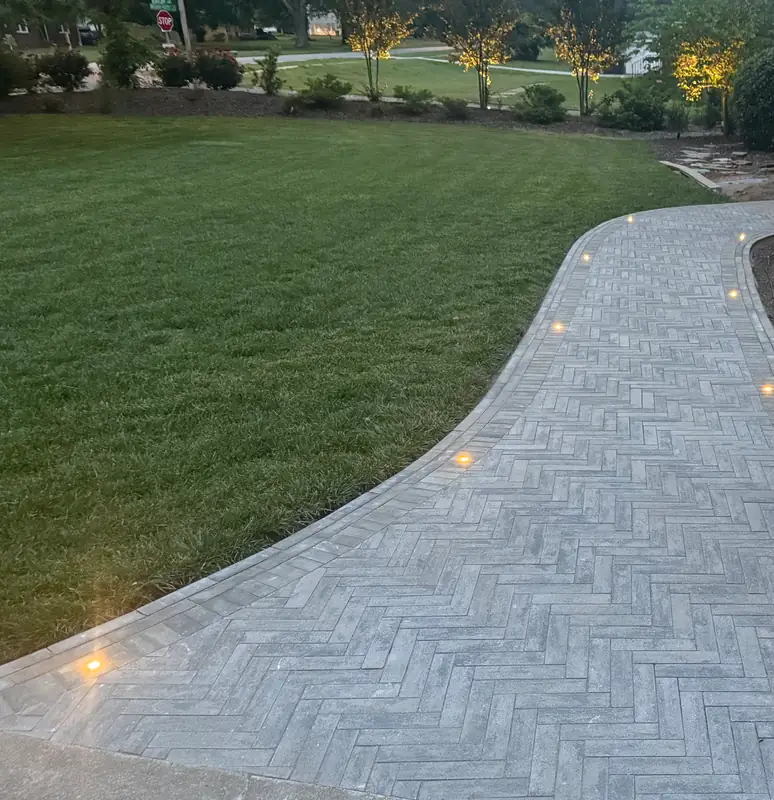 Herringbone paver walkway curving across a lawn, lit by in-ground landscape lights at dusk